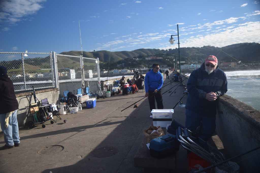 People fishing along the railing of the Pacifica pier with coastal hills in the background