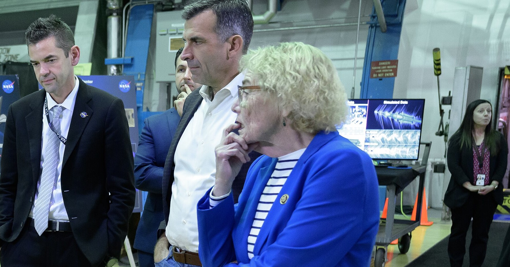 Rep. Zoe Lofgren, in a blue jacket, listens during a briefing at NASA Ames Research Center, with Rep. Sam Liccardo and NASA Administrator Jared Isaacman standing beside her.