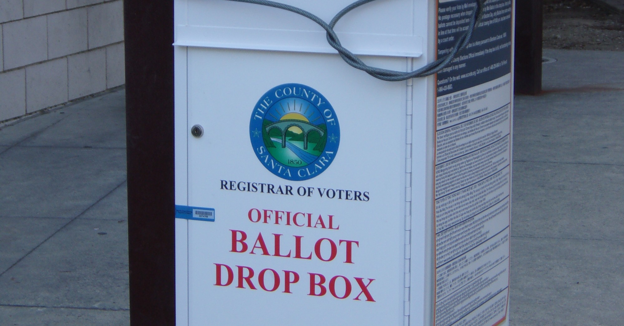A white Santa Clara County Registrar of Voters official ballot drop box with red lettering, outside a San Jose library
