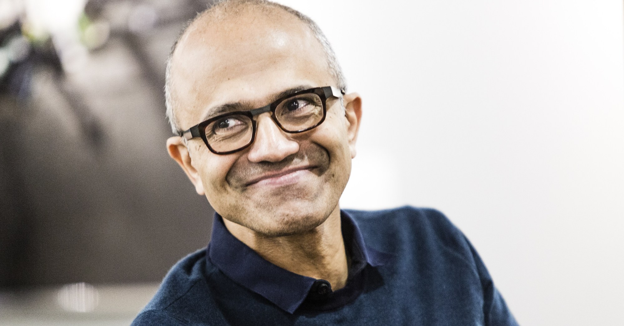 Microsoft Chairman and CEO Satya Nadella, wearing glasses and a dark sweater, smiles in a portrait.