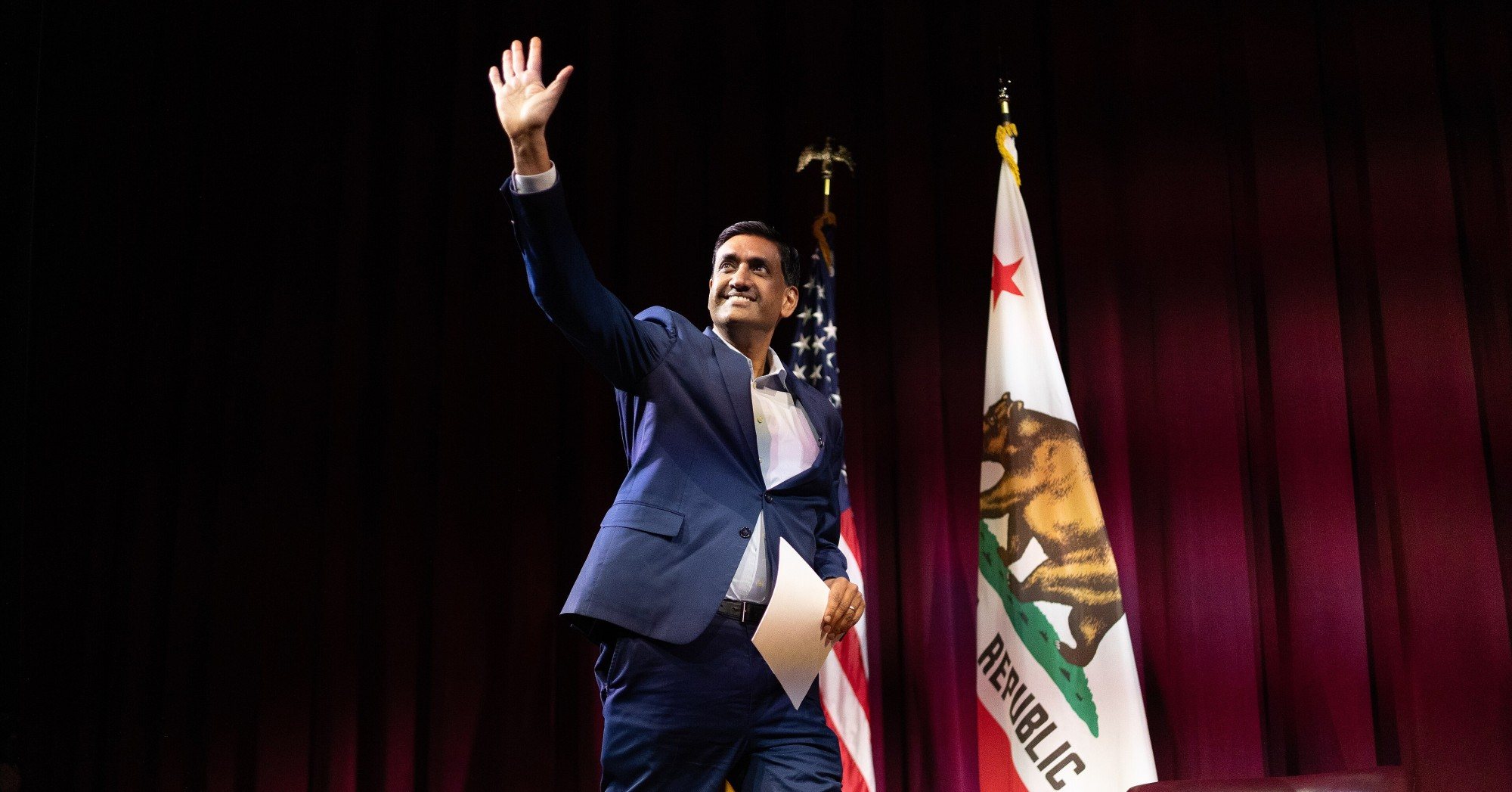 Rep. Ro Khanna waving on stage in front of U.S. and California flags
