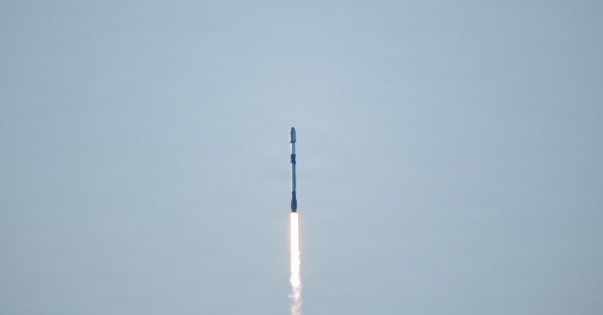 A SpaceX Falcon 9 rocket climbs into a pale gray sky trailing a bright exhaust plume after launch from Vandenberg Space Force Base